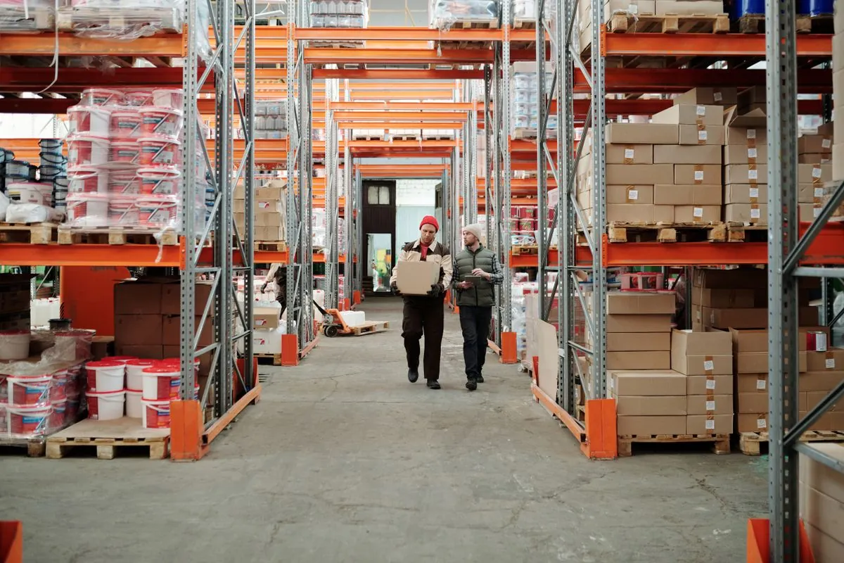 Two people walking between stocked pallet racks in a distribution warehouse.
